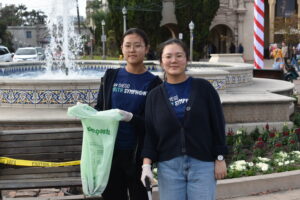 Two women wearing blue shirts, each holding a green bag, sharing a cheerful moment outdoors.