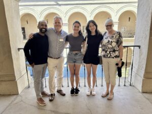 A group photo featuring KeyNote’s 2024–2025 Youth Board Members, Madeleine and Elizabeth, standing together with their adult board mentor, KeyNote’s Artistic Director, and KeyNote’s CEO. All are smiling and standing against a neutral background overlooking Balboa Park's Casa del Prado building.
